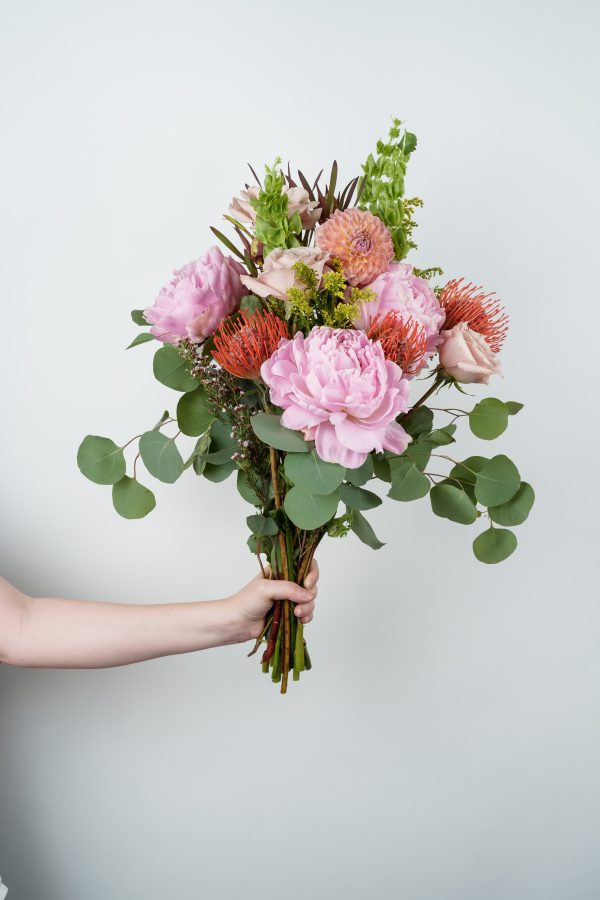 Hand holding a modern bouquet with pink peonies, roses, pincushion protea, and eucalyptus against a white background