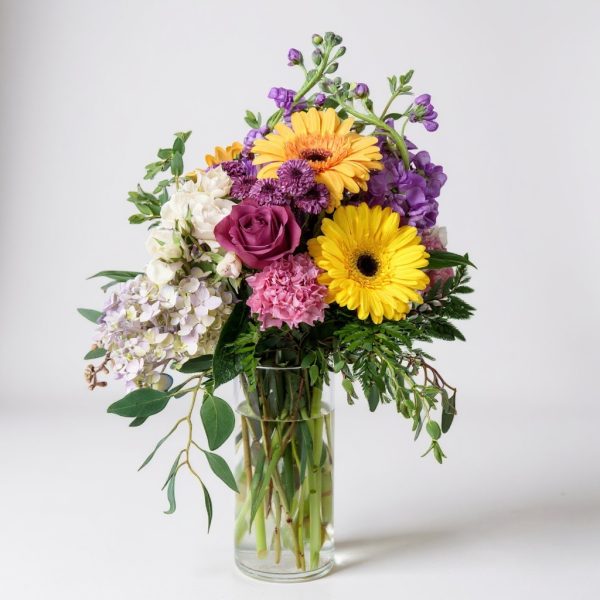 Tall yellow and purple mixed flower arrangement with gerbera daisies, purple roses, carnations, hydrangea, and stock in a clear cylinder vase