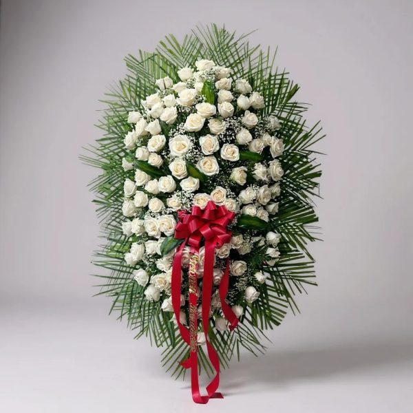 Funeral standing spray with white roses, baby’s breath, greenery, and red ribbon bow on easel