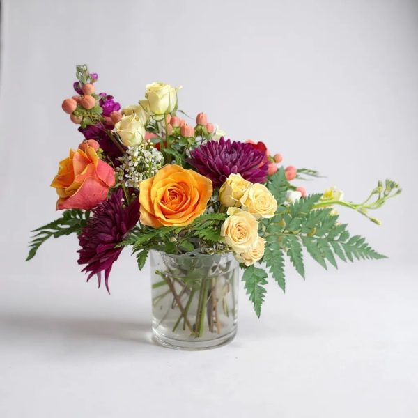 Bouquet with orange roses, yellow spray roses, burgundy mums, and greenery in a glass vase