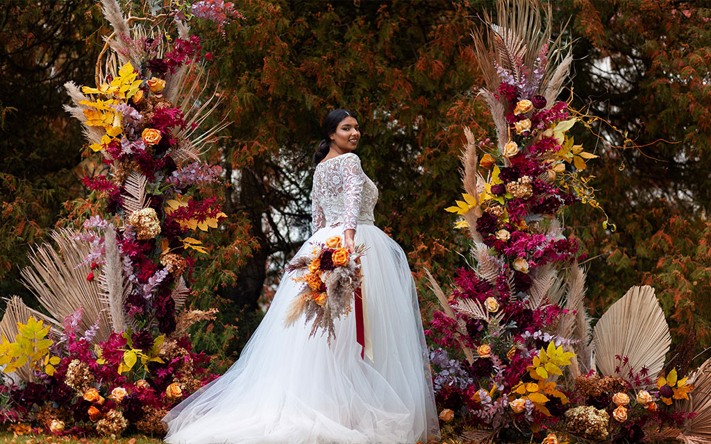 Fall wedding flowers ceremony arch with tall pampas grass, dried palm fronds, burgundy roses, orange dahlias, and autumn leaves
