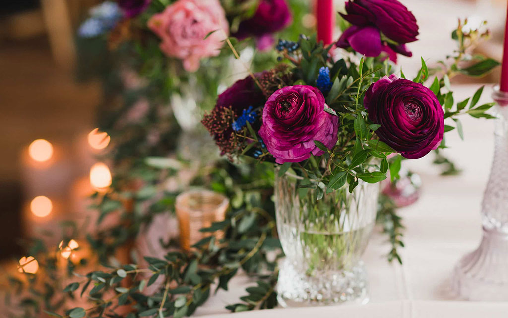 Seasonal wedding flowers centerpiece with magenta ranunculus and blue accents in crystal vase with candlelight for spring reception