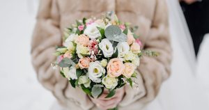 Winter wedding flowers bridal bouquet with white and peach roses, lisianthus, and eucalyptus held by bride in elegant winter setting